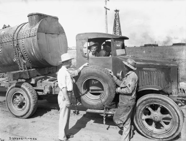 Men standing next to an International heavy-duty oil truck. Two delivery drivers from Barrington Transfer Company are in the cab of the truck. An oil well is in the far background.