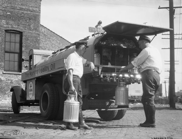 Two men are filingl oil cans from the back of an International truck operated by Mobiloil. Text on the side of the truck reads: "The World's Quality Oil" and "Mobiloil Certified Service."