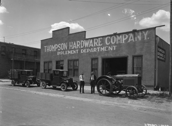 IH Trucks and Tractor in front of Thompson Hardware Co. Two men are standing in the street between two IH trucks and one IH tractor in front of the "Thompson Hardware Company Implement Department," most likely an International Harvester dealership. A sign on the side wall of the building reads: "Thompson Hardware Co Implements."