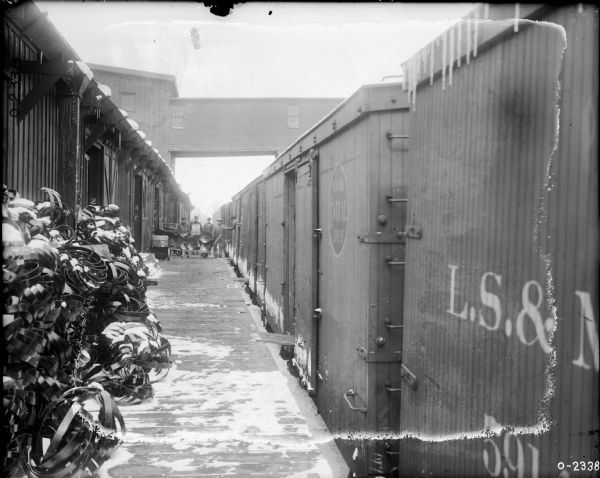 View down platform of train stopped at a loading dock at International Harvester's Osborne Works (later known as Auburn Works). On the dock is a large stack of scrap metal or parts. Further down the dock four men wearing work clothes stand near a cart, and the factory building is high above them in the background. There is snow on the ground and icicles hanging from the train.