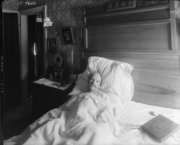 A man is lying in bed, covered to his chin in bedding and propped up by pillows. On the right side of the bed is a large bible and a rosary. The room is richly decorated with a large wooden headboard and wallpaper. Beside the man is a table with tablecloth on which is sitting a religious shrine. There is also a glass candle holder in the shape of a cross, what appears to be an ink well and pen, and another jar. Above the table are two religious paintings and a lamp with a card attached to the side of the glass shade. This man may have been an employee at International Harvester's Osborne Works in Auburn, New York.