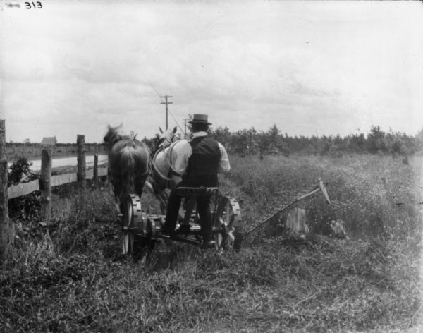 Rear View of Man on Mower Rear view of a man on horse-drawn mower. There is a fence and a road on the left. The mower blade is partially lifted to pass over a tree stump.