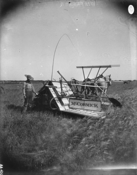 Man Standing in Field with Horse-Drawn Binder Man standing near the back of a horse-drawn McCormick binder in a field. The two horses stand waiting while the man appears to be using an oil can.