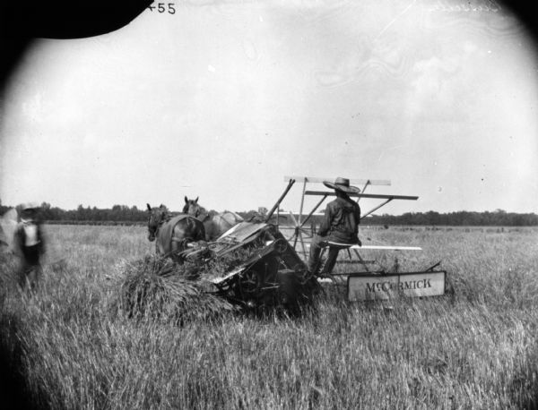 Men Using Horse-Drawn Binder in Field Three-quarter rear view from left of a man using a McCormick horse-drawn binder in a field. There is another man standing on the left.