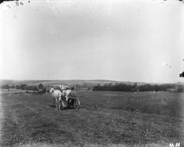 Man Using Horse-Drawn Mower in Field View from rear of a man using a horse-drawn mower in a field. Fences, trees and hills are in the background.