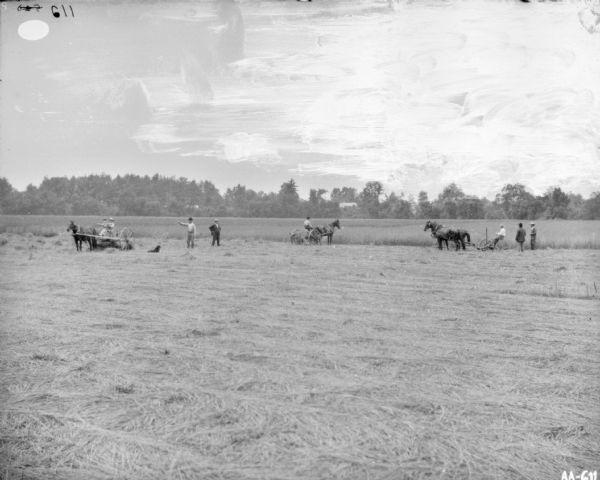 Men Using Horse-Drawn Mowers in Field View across partially harvested field towards a group of people posing. On the left are what may be young children posing on a horse-drawn vehicle. A man is standing with an arm raised towards what may be a dog sitting in the field. Next to them another man is standing looking towards a man on a horse-drawn cart or agricultural machinery. On the right two men are standing near a man operating a horse-drawn mower.