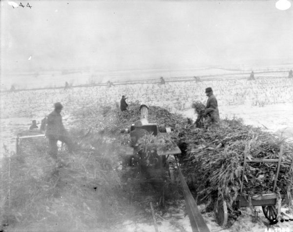 Silaging Operation Slightly elevated view of a silaging operation. The husker and shredders are belt-driven with the belt is in the foreground. Men are on wagons piled with silage on each side of the husker and shredder, which another man is operating. Two other men are standing in the snowy field behind.