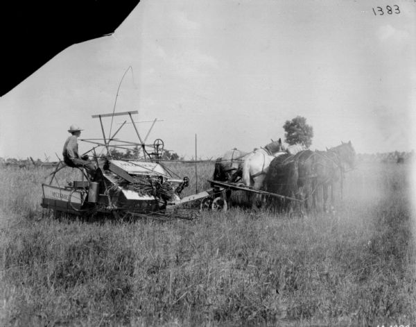 Man Using Horse-Drawn Binder in Field Right side view of a man using a team of four horses on a horse-drawn McCormick binder in a field. Two of the horses are wearing fly-nets. A split-rail fence is in the background.