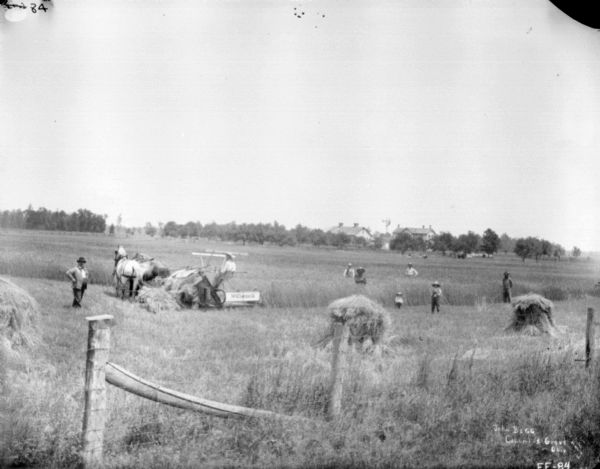 Group of People at John Begg Farm View over fence across field towards group of people standing near a man on a horse-drawn McCormick binder. Farm buildings are in the far background.
