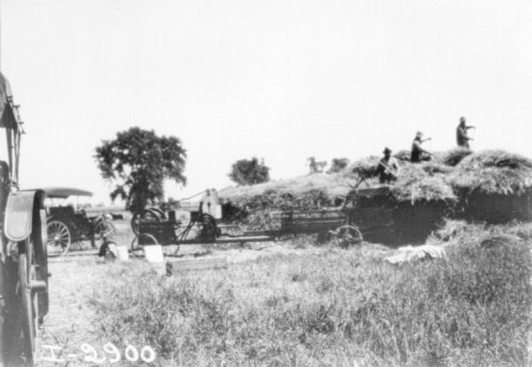 Hay Presses with Large Stack of Hay Men are working with hay presses near a large stack of hay in a field. There is a tractor in the left foreground.