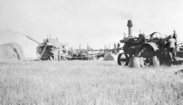 Harvesting Operation View across field towards a large harvesting operation. On the far left is a large haystack. The thresher is belt-driven by a tractor in the right foreground. A large group of men are working near the thresher, and a horse-drawn wagon is in the center.