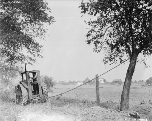 Tractor Pulling Down Tree View from rear of a man using a tractor and chain to pull down a tree to clear a field. Farm buildings are across a field in the far background.
