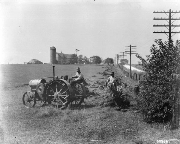Men Pulling Tree Stump with Titan Tractor Two men are pulling tree stumps in a field with a Titan tractor. A silo and farm buildings are in the background.