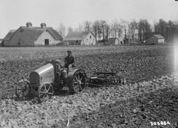 Man Disking a Field with Tractor Three-quarter view from front left of a man disking a field with a tractor. There are farm buildings in the background.