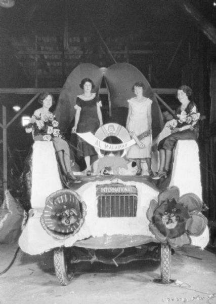 Women Posing on International Harvester Float Group portrait of women wearing gowns posing on an International Harvester float. They are sitting and standing on the front of the decorated vehicle, along with a sign for: "Al Malaikah."