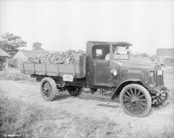 Coal Delivery Truck Three-quarter view from front right of a man sitting in the driver's seat of a coal delivery truck parked on a dirt road. A sign on the side of the truck bed reads: "Happy Coal."
