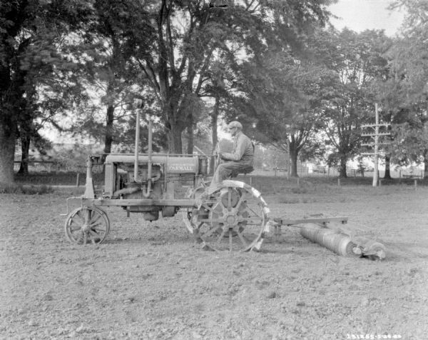 Left side profile view of a man driving a Farmall pulling a cultivator in a field.