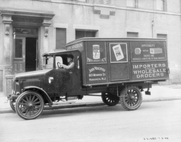 View across street towards a man sitting in the driver's seat of a truck parked along a curb. There is an entrance to a building in the background. Signs painted on the truck read: "John Minervini, Importers and Wholesale Grocers."