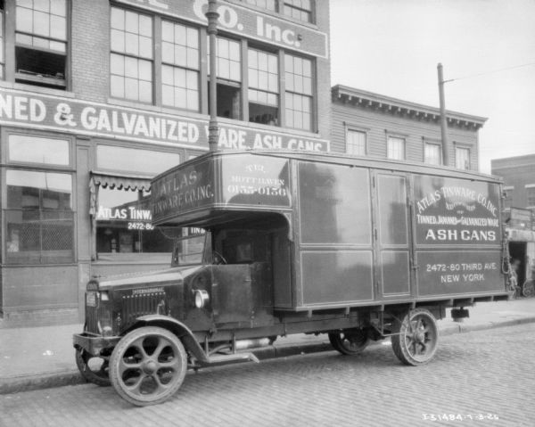 View across street towards a truck parked along the curb in front of a brick building. Signs on the truck and the building read: "Atlas Tinware Co. Inc. Manufacturers of Tinned, Japanned and Galvanized Ware, Ash Cans."
