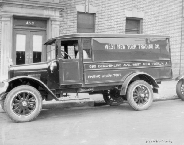 View across street towards a man sitting in the driver's seat of a truck parked along the curb in front of a brick building. The sign on the side of the truck reads: "West New York Trading Co."