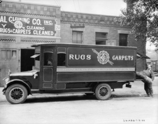View across street towards a man loading a rug into the back of a delivery truck. There is a man sitting in the driver's seat. The building in the background has a sign on the front that reads: "Cleaning Co. Inc." The sign on the truck reads: "General Cleaning Co. Inc., Rugs, Carpets."