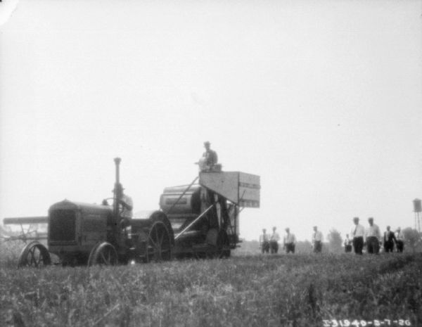 Thresher Operation in Field View across field towards demo of new machine(?) with a crowd of well-dressed men observing.