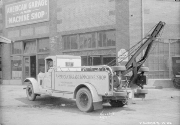 Three-quarter view from rear left of a tow truck for American Garage & Machine Shop. There is a brick building in the background, which also has a sign above the entrance for "American Garage and Machine Shop."