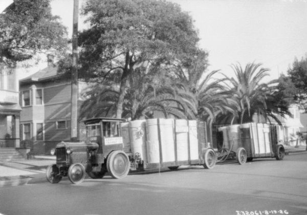 View across street towards an industrial tractor pulling two trailers. There are buildings along the curb in the background. A sign on the tractor reads: "Associated Transit Co."