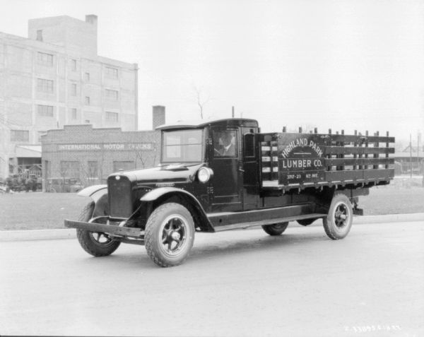 Lumber Delivery Truck Three-quarter view from front of the driver's side of a lumber delivery truck. A man is sitting in the driver's seat, and a sign on the side of the stake body reads: "Highland Park Lumber Co." A building in the background has a sign on the front that reads: "International Motor Trucks."