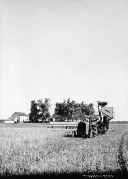 Man Driving Tractor Pulling Harvester Thresher View across field towards a man driving a tractor. The tractor is pulling a harvester thresher, and another man is sitting on the front of the thresher. Farm buildings are in the background among trees.