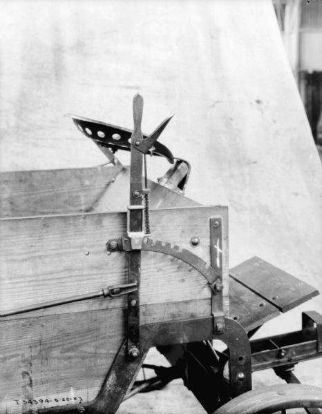 Close-up of seat, footboard, and handle of control lever at the front of a manure spreader. There is a backdrop in the background in what may be a factory.