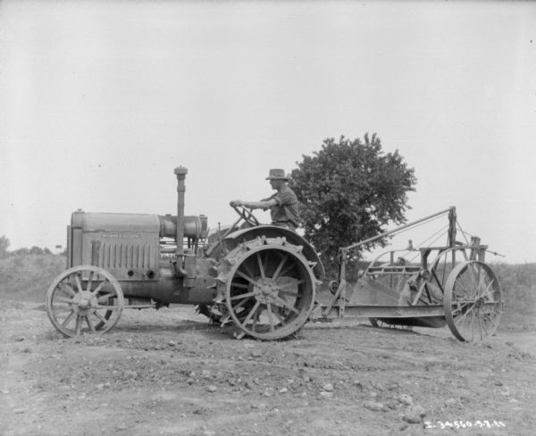 Man Driving Industrial Tractor Left side profile view of a man driving a McCormick-Deering industrial tractor which is pulling an implement on a trailer with large wheels over bare dirt ground.