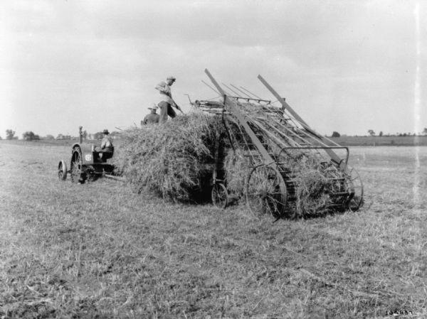 Hay Loaders Two men are working on top of a pile of hay to put into a hay loader. In the background is a man operating a tractor.