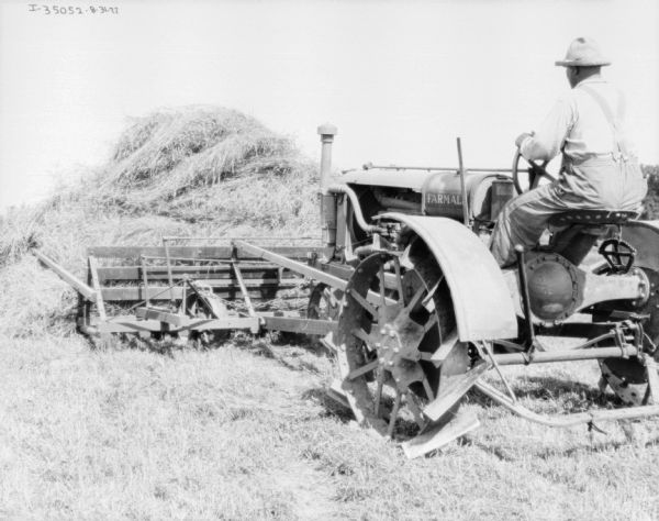 Man Moving Hay with Farmall Tractor Three-quarter view from left rear of a man using a tractor to move a pile of hay in a field.