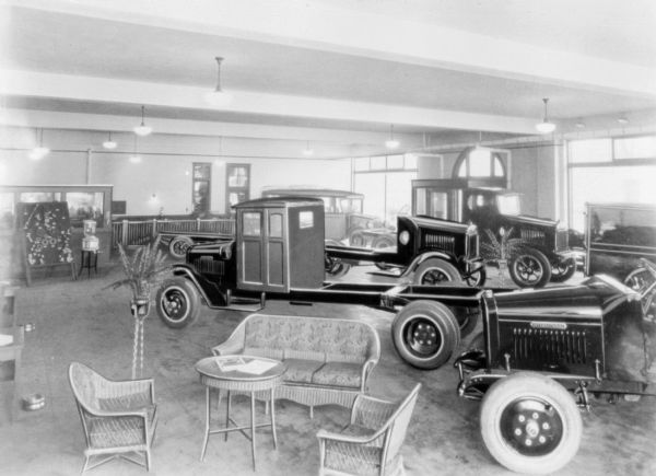 Trucks are parked in a showroom. In the foreground is a couch, chairs and a table. Machine parts are displayed on a board near a water cooler in the background.