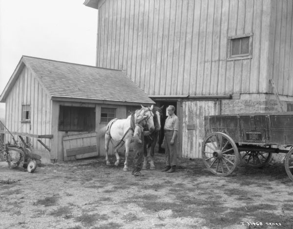 Man and Boy with Two Horses A man and boy are standing with two horses in front of a barn and small building. On the right is a McCormick-Deering Weber wagon.