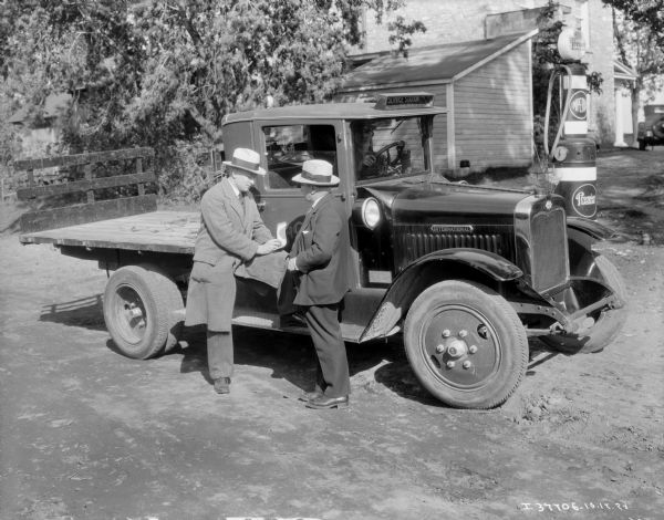 Two Men with International Truck for Service Station Two men are standing alongside the running board of the passenger side of an International truck. The truck has an open bed, with a gate at the front and back. A man is sitting in the driver's seat of the truck, which has a sign on the roof that reads: "Service Station SH.5169." A group of five small United States and United Kingdom flags are attached to the front of the truck hood, above the Triple Diamond International logo. Behind the truck is a Premier Gasoline pump, and there are buildings and an automobile in the background. There is a Quebec license plate on the front of the truck.