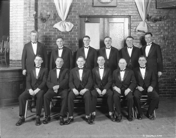 Group Portrait of Men Formal indoor group portrait of six men sitting, and six men standing. They are all wearing suits and bow ties. There is a stage on the left.