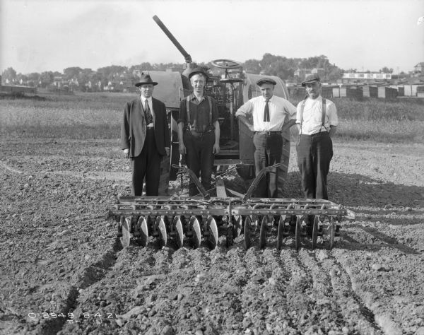 Four Men Posing with Tractor Hitched to Disc Harrow Group portrait of four men standing between a disc harrow and a tractor in a field. The disc harrow is in the foreground, and the tractor is behind them. The man on the far left is wearing a suit and a hat. The other three men are wearing work clothes and hats. In the far background are stacks of lumber, and in the far background are houses on a hill among trees.
