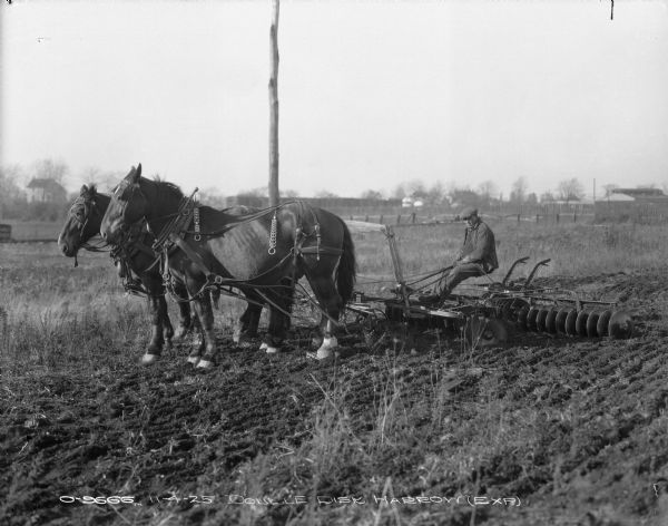 Horse-Drawn Double Disk Harrow Three-quarter view from front left of a man sitting on a double disk harrow (experimental) being pulled through a field by four horses. A fence and buildings are in the background.