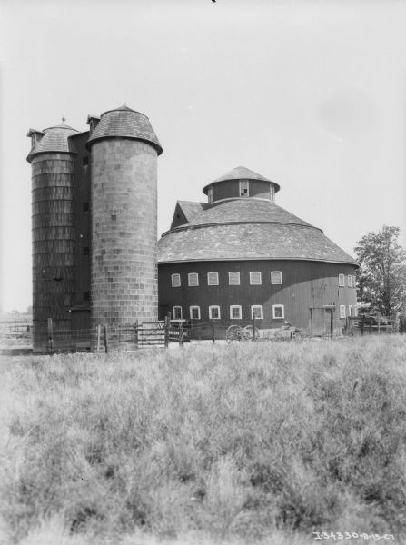 View across field towards two silo's next to a round barn. There is a wagon behind a fence just in front of the barn, and a horse is standing inside a fence on the far right.