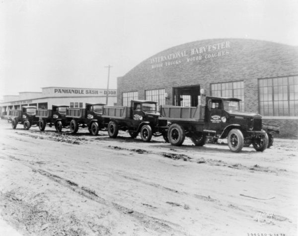 View towards five delivery trucks parked in a long row. The sign painted on the passenger side door of one of the trucks reads: "Barker Bro's." In the background is a brick building with a sign that reads: "International Harvester, Motor Trucks, Motor Coaches." Another building in the background on the left has a sign for "Panhandle Sash and Door."