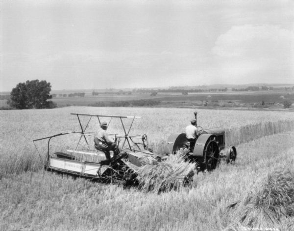 Tractor Drawn Binder Slightly elevated view from right rear towards a man driving a tractor in a field. The tractor is pulling another man who is sitting on a binder.