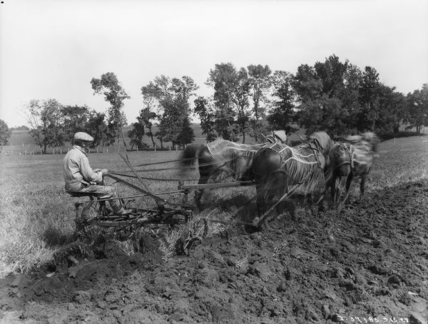 Man on Horse-Drawn Plow Right side profile view of a man using a team of four horses to pull a McCormick-Deering P&O plow in a field. The horses are wearing fly-nets.