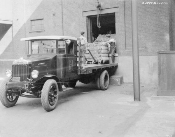 Truck at Loading Dock View towards a truck backed up at a loading dock in a brick building. Men are using a cart to move a large load of material. There is a pulley hanging above the door.