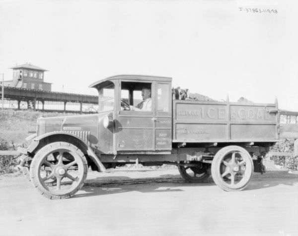 Man Sitting in Delivery Truck View towards a man sitting in the driver's seat of a delivery truck. The sign painted on the truck reads: "Hollywood Ice & Coal." In the background is a building, and a bridge or elevated railway.