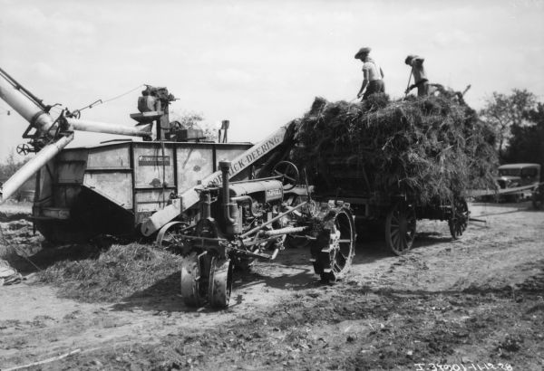 Farmall Parked at Threshing Operation A Farmall tractor is parked in front of a wagon and a thresher. Two men are standing on top of hay stacked on the wagon. He is feeding the McCormick-Deering thresher. An automobile is parked in the background on the right.