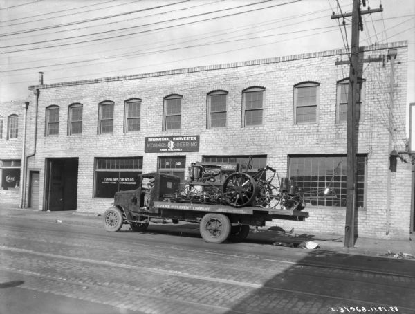 View across street towards a man sitting in the driver's seat of a truck.  On the truck bed is a Farmall trator and lister. The truck is parked in front of a brick building, which has a sign for Evans Implement Co. in the window.