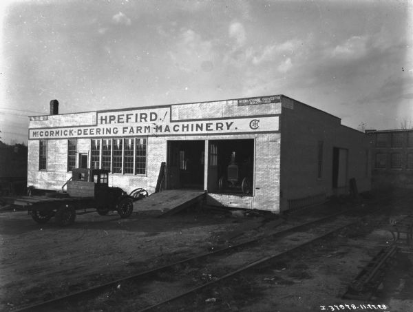 Exterior view across railroad tracks towards a brick building. A truck is parked at the base of the ramp leading into the repair shop. A tractor is sitting inside the other open garage door. The sign painted on the front reads: "H.P. EFIRD.! McCormick-Deering Farm Machinery."