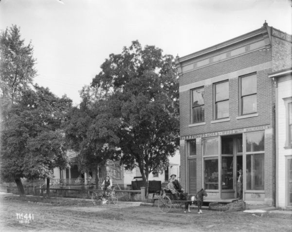 View across street towards a man on a buggy on the curb at the left, and a man and boy on another buggy being pulled by a pony or miniature horse. They are near a storefront with a sign that reads: "E.P. Phillips, Coal-Wood & ? Machinery." People are sitting on a porch in the background on the left.